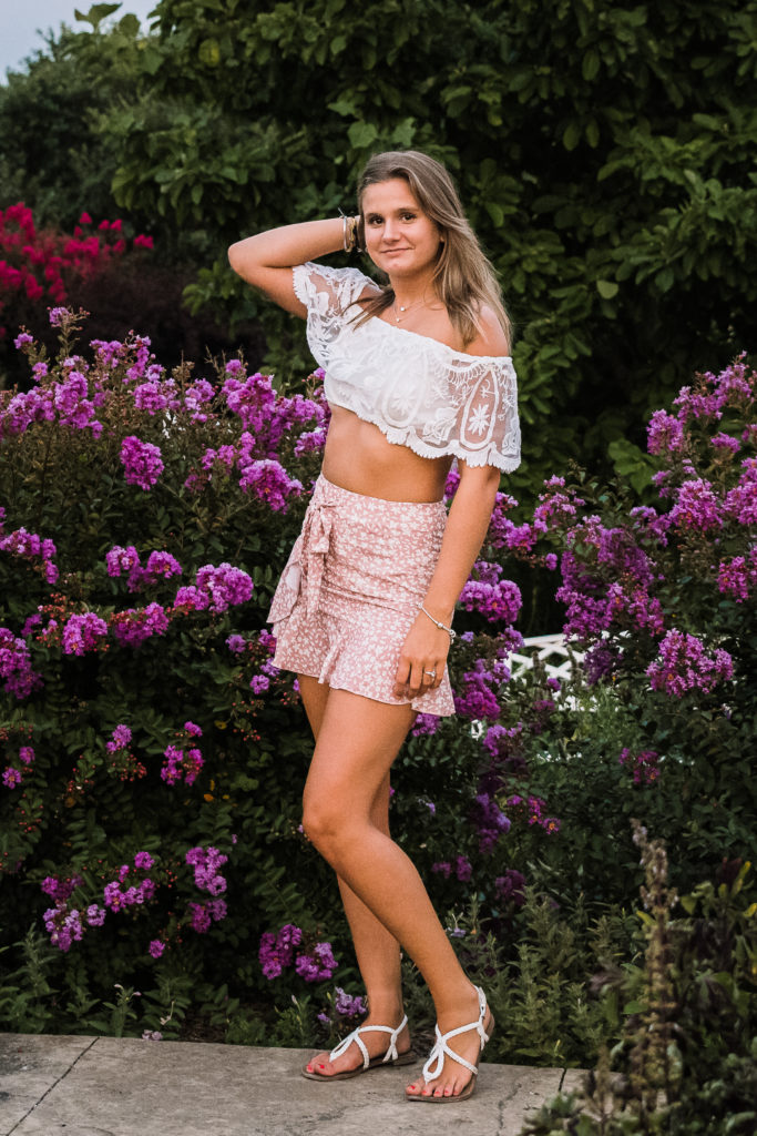 senior girl standing in flower garden with white crop top and pick skirt placing hand gently on hair