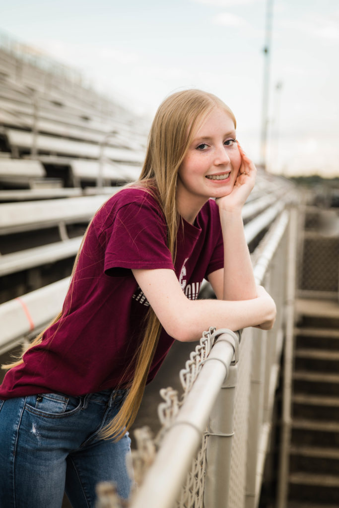senior girl in football stadium with hand under chin leaning against fence