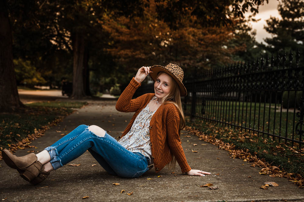 Senior girl sitting on ground holding one hand on leopard print hat with other hand resting on ground