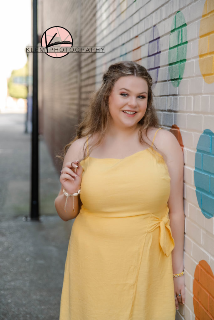 Summer senior pictures evansville indiana by klem photography. senior in yellow knee length dress leaning up against a brick wall downtown and smiling