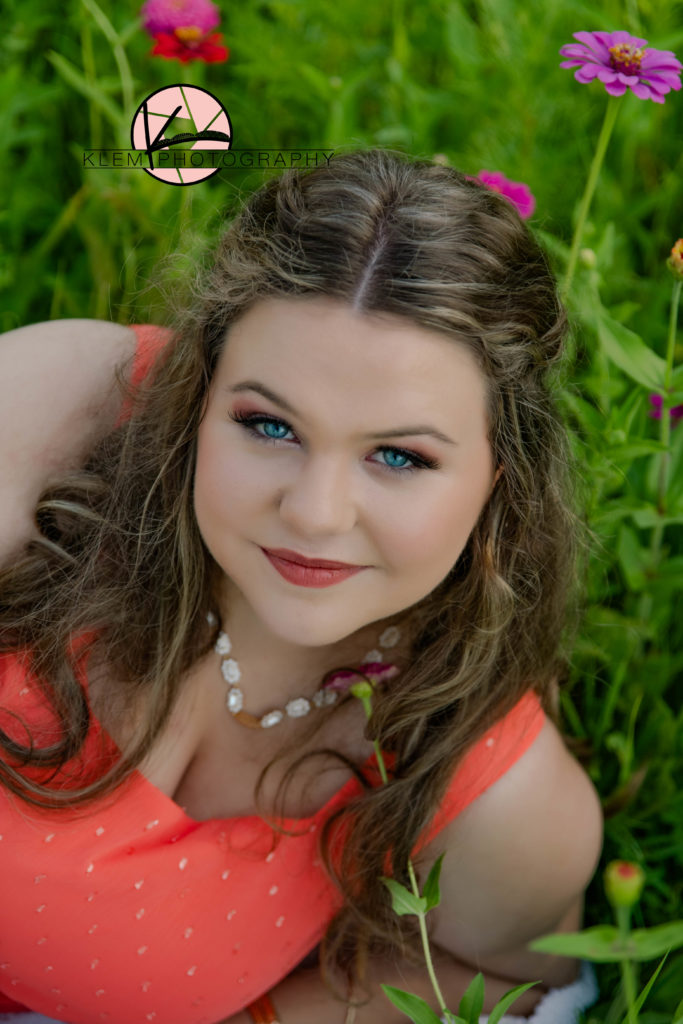 Summer senior pictures evansville indiana by klem photography. senior in coral maxi dress laying in a field of wildflowers smiling with bold blue eyes