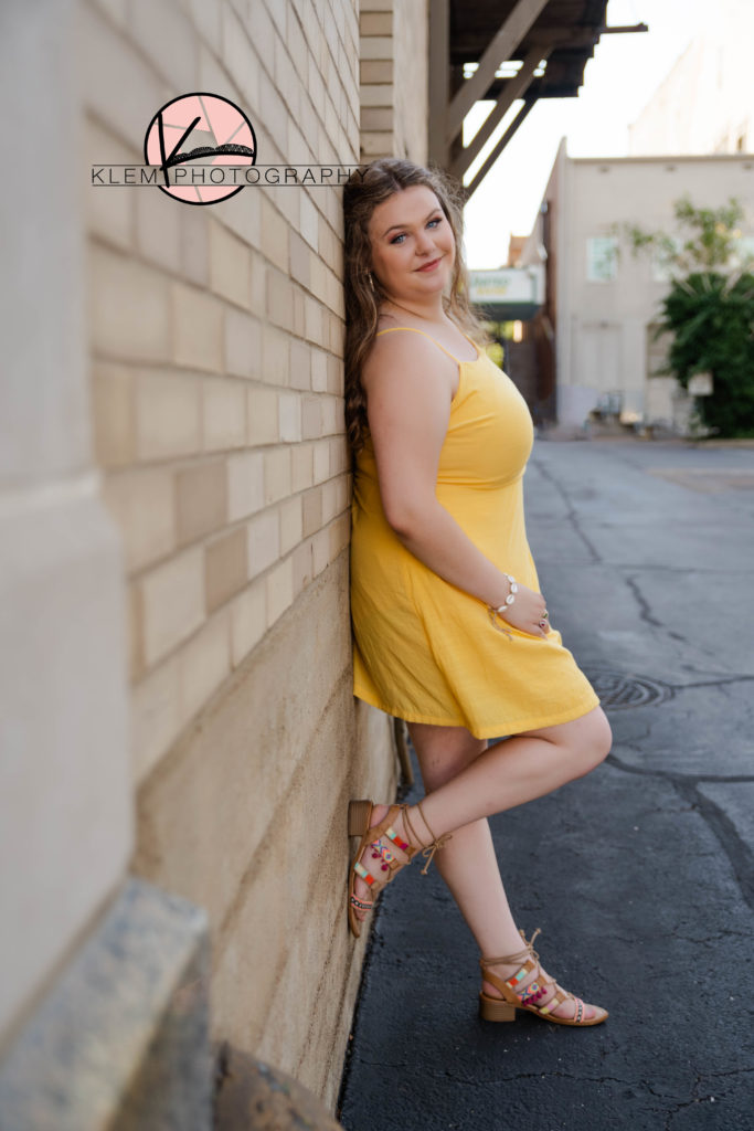 Summer senior pictures evansville indiana by klem photography. senior in yellow knee length dress with strappy bold sandals leaning up against a brick wall downtown and smiling
