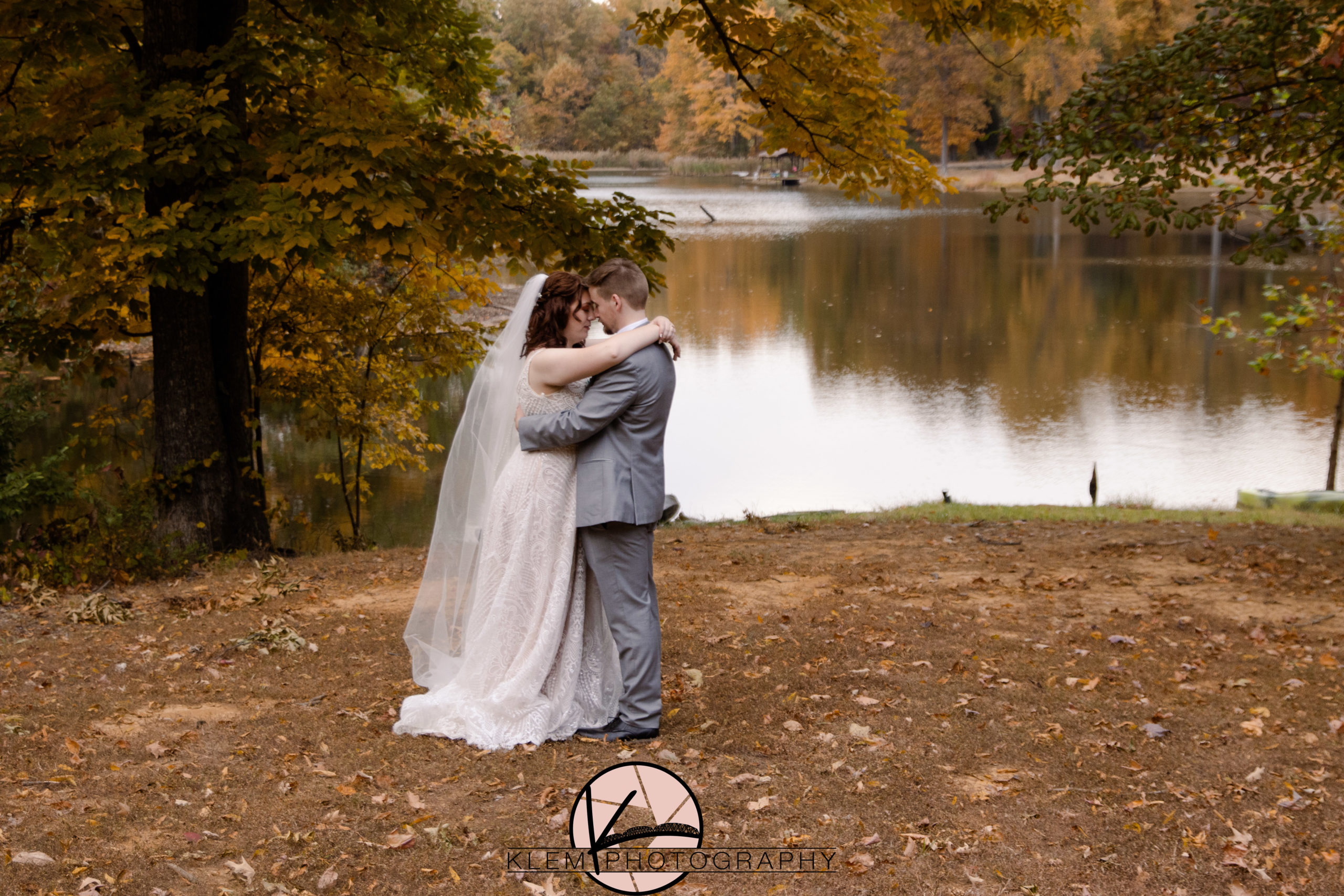 Bride and groom stand in front of lake with brides arms wrapped around grooms neck and his hands at her waist as they look into each others eyes and the fall colors surround them at their fall wedding in kentucky