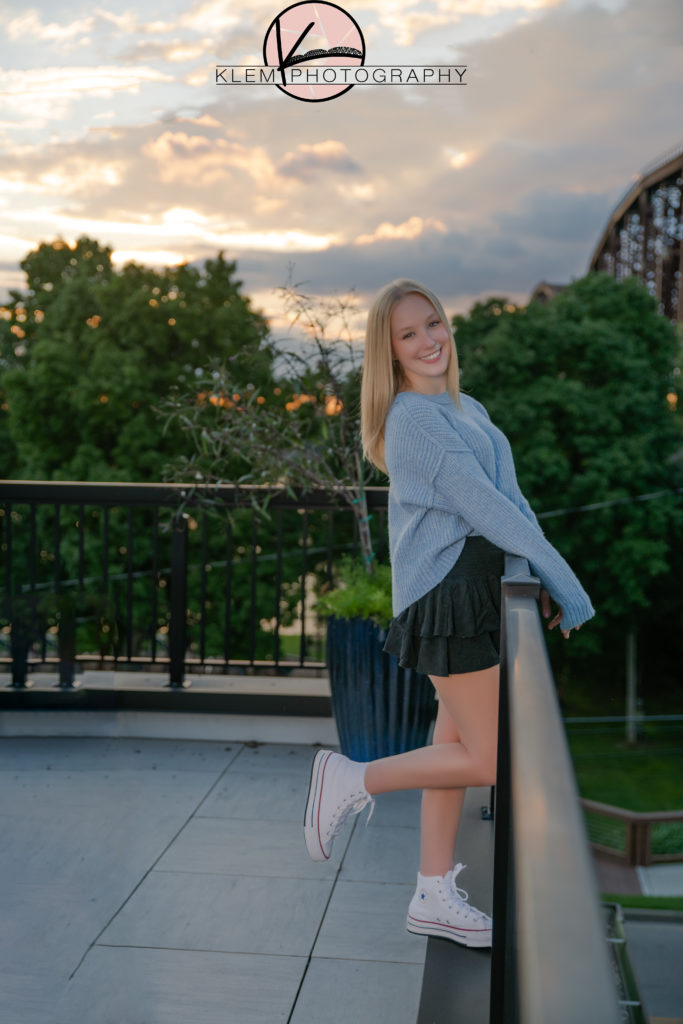 kentucky senior pictures taken on rooftop in henderson kentucky as senior girl with blonde hair stands with one leg kicked back behind her in blue sweater and black skirt smiling at camera while a beautiful sunset is in the background. 