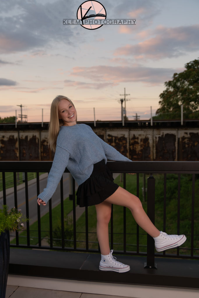 kentucky senior pictures taken on rooftop in henderson kentucky as senior girl with blonde hair stands with one leg kicked in front of her in blue sweater and black skirt smiling at camera while a beautiful sunset is in the background. 