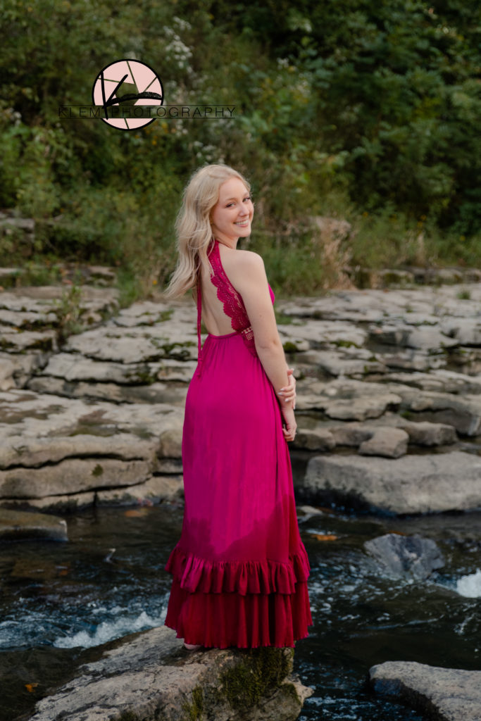 Senior girl in pink maxi dress stands on rock in waterfall and looks at the camera smiling for a senior picture from kentucky senior pictures by klem photography 