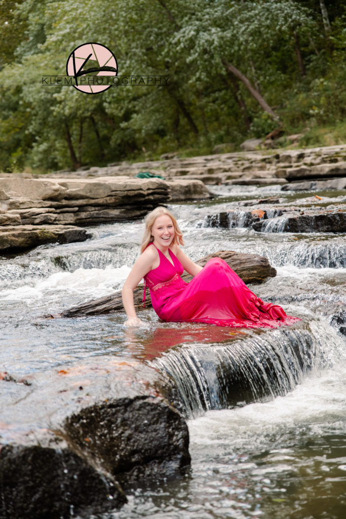 Senior girl in pink maxi dress sits on waterfall and looks at the camera laughing for a candid senior picture from kentucky senior pictures by klem photography 