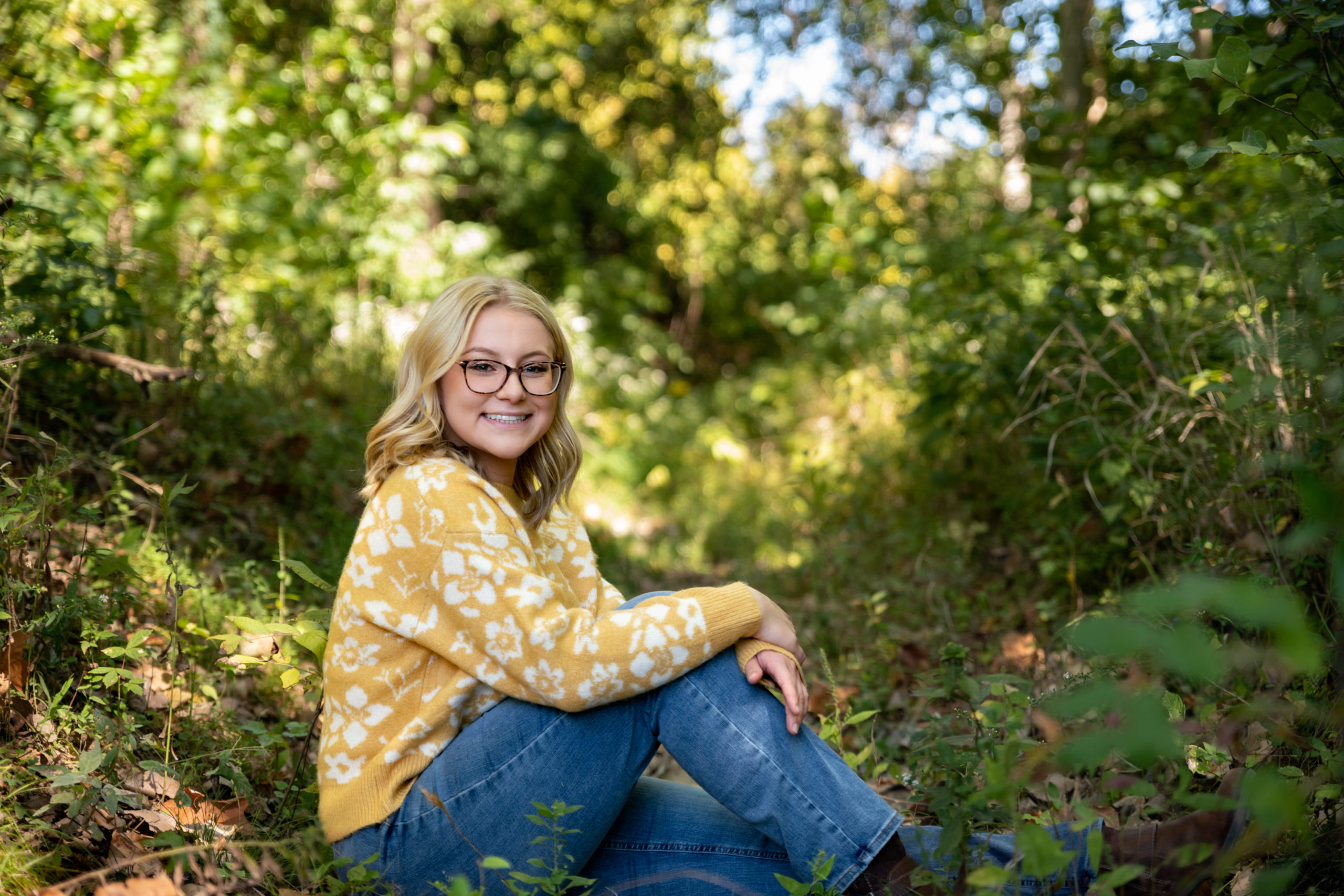 fall senior pictures in henderson ky as senior girl with blonde hair and glassessits on the ground of the woods wearing a mustard colored sweater, denim blue jeans and brown booties creating the perfect fall look