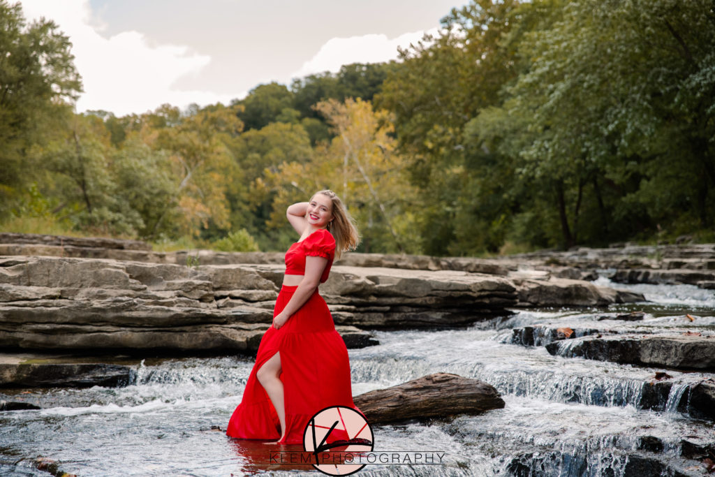 senior pictures in kentucky with senior girl in bold red dress standing in the waterfalls as her dress blows in the breeze