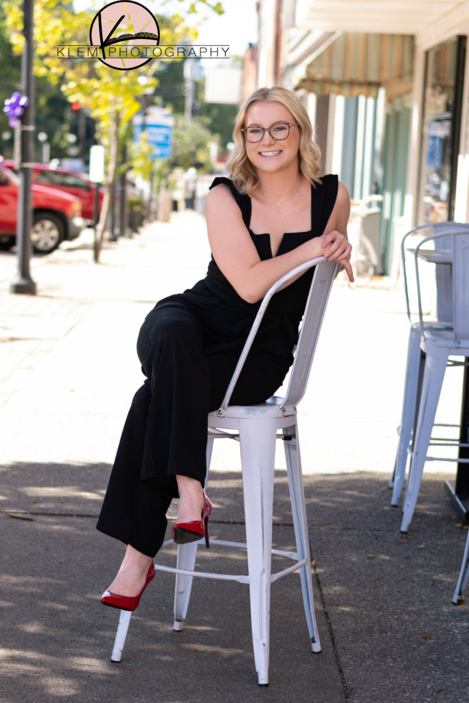 High school senior girl sits on tall chair on sidewalk wearing black jumpsuit and bold red high heels by kentucky senior photographer klem photography 