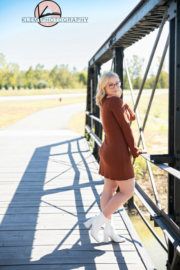 senior pictures henderson ky in the fall, senior girl wearing a burnt orange dress with white booties standing on a steel bridge with hand on shoulder by klem photography kentucky senior photographer
