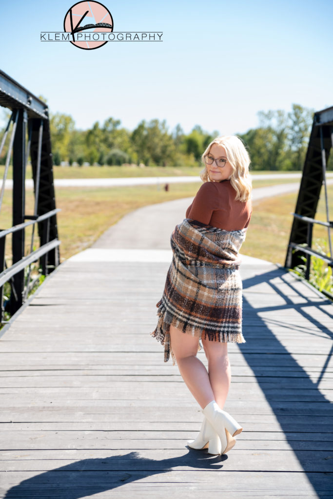 senior pictures henderson ky in the fall, senior girl wearing a burnt orange dress, plaid shawl, and white booties standing on a steel bridge looking over her shoulder by klem photography kentucky senior photographer