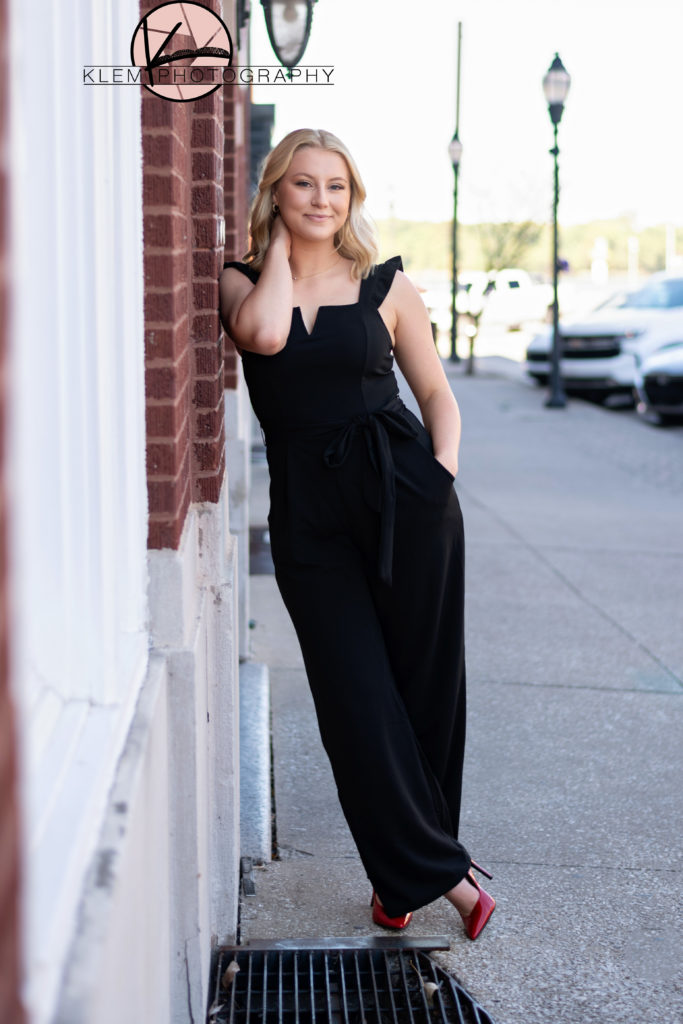 High school senior girl leans on wall on sidewalk wearing black jumpsuit and bold red high heels by kentucky senior photographer klem photography 