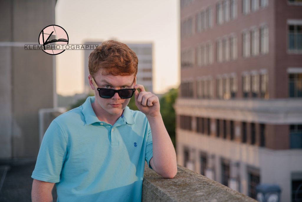 guy senior session in evansville indiana on rooftop at 3rd street parking garage by klem photography kentucky senior photographer. guy is wearing blue polo shirt and sunglasses while he stares at the camera. He has wavy red hair