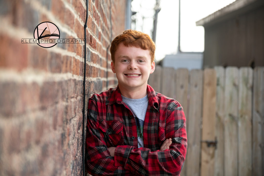 guy senior pictures by kentucky senior photographer klem photography guy with red hair leaning against brick wall. he is wearing a plaid button down shirt and he is smiling at the camera
