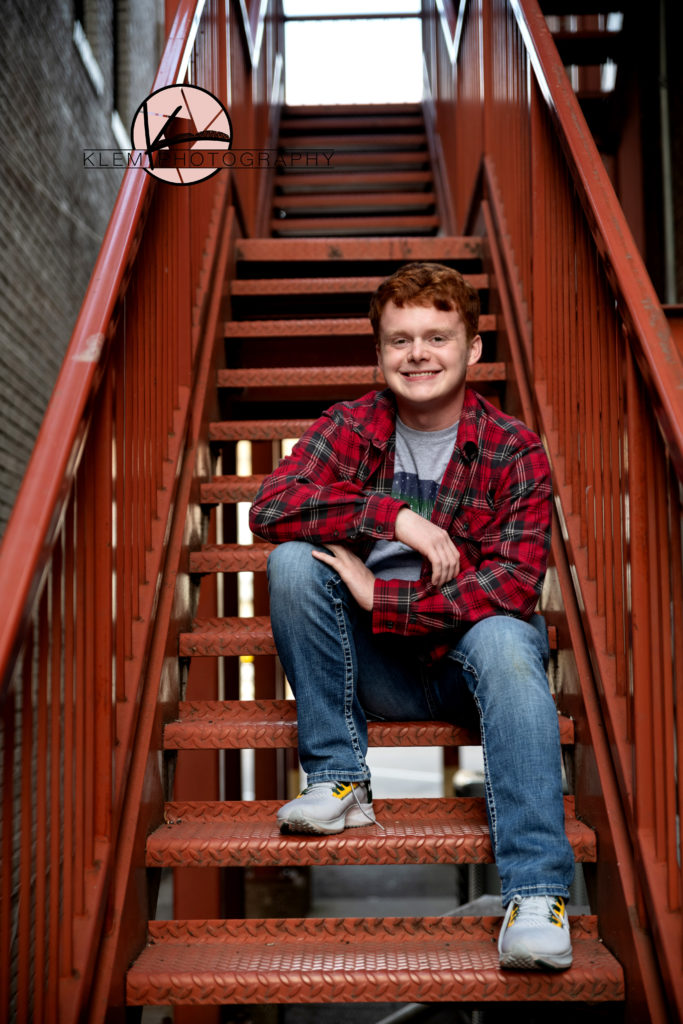 guy senior session in kentucky by klem photography kentucky senior photographer. guy is wearing red plaid shirt and jeans. he is sitting on some rusty stairs. He has wavy red hair