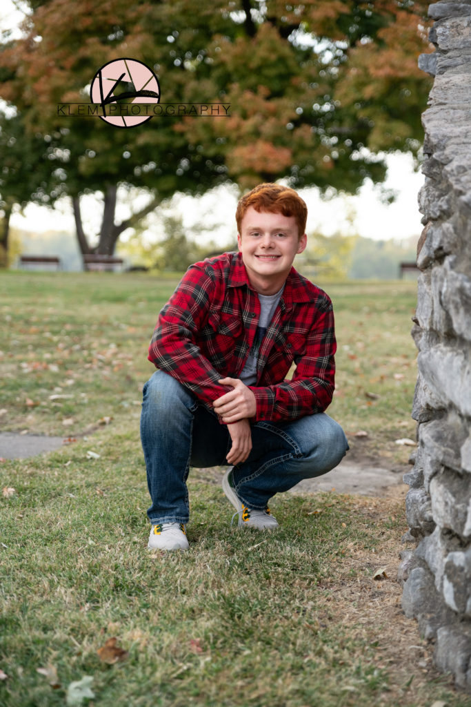 guy senior session in kentucky by klem photography kentucky senior photographer. guy is wearing red plaid shirt and jeans. He is kneeling on one knee. He has wavy red hair