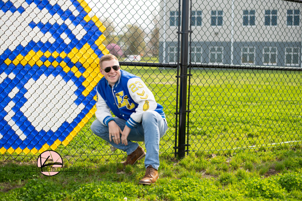 evansville senior photos by klem photography at evansville christian school. senior boy is squatting down in front of a chain link fence. he is wearing a blue and white letterman jacket and blue jeans