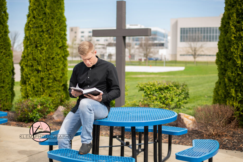 evansville senior photos by klem photography at evansville christian school. senior boy sits on blue table in front of cross reading his bible. he is wearing a black shirt and blue jeans
