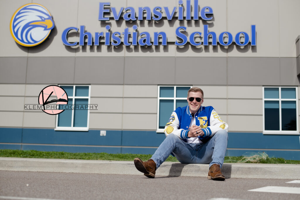 evansville senior photos by klem photography at evansville christian school. senior boy is sitting in front of the evansville christian school sign wearing a blue and white letterman jacket and blue jeans