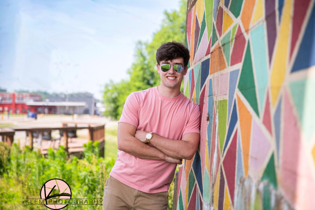 senior boy wearing a pink shirt and sunglasses leaning up against a painted wall mural in downtown henderson kentucky with his arms crossed