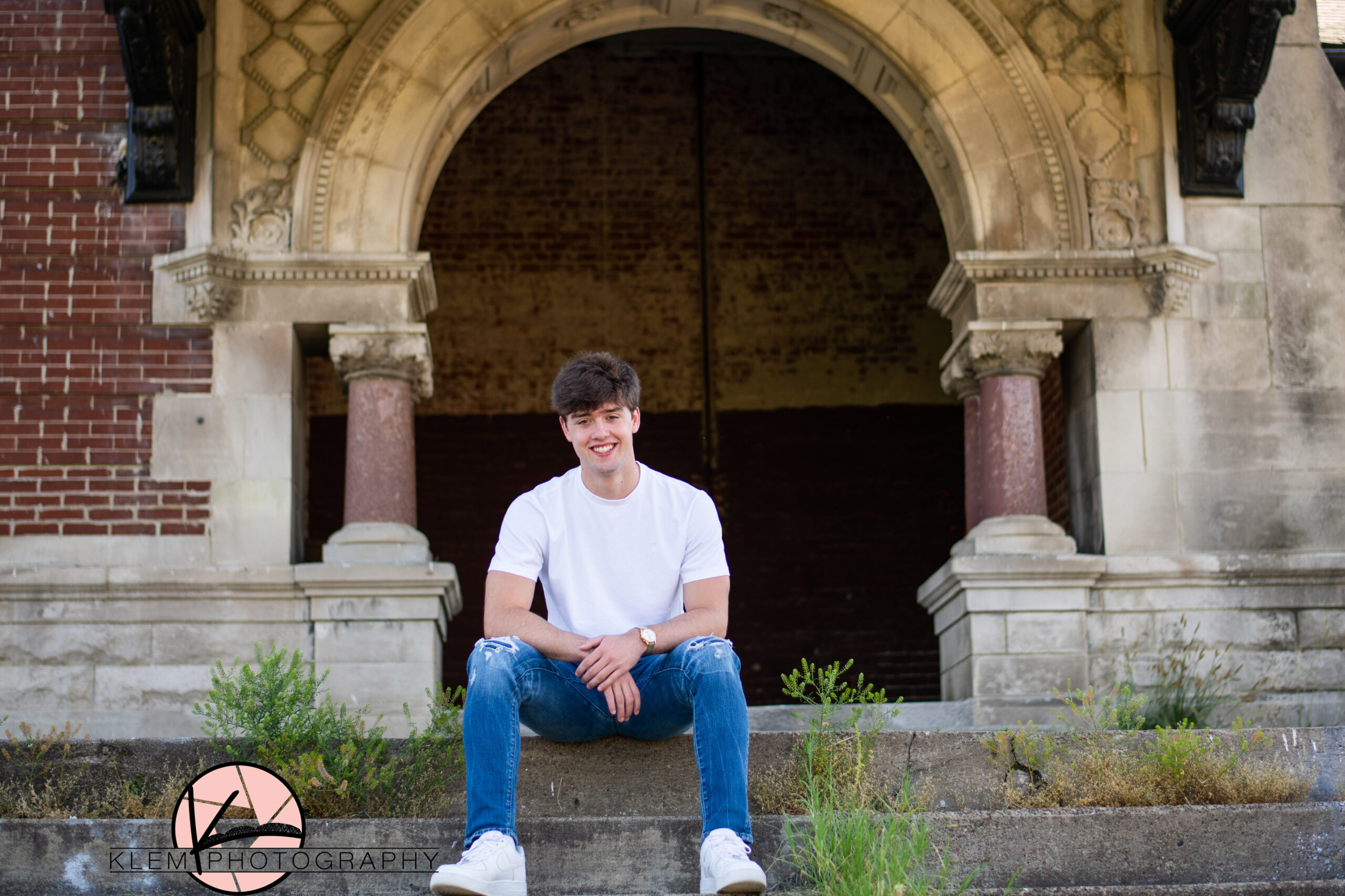 senior boy wearing a white shirt and blue jeans sits in front of old train depot in henderson kentucky for an urban senior picture setting
