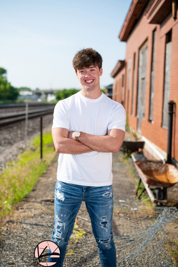 urban senior pictures in henderson kentucky senior boy stands with arms crossed and smiling. he is wearing a white shirt and blue jeans
