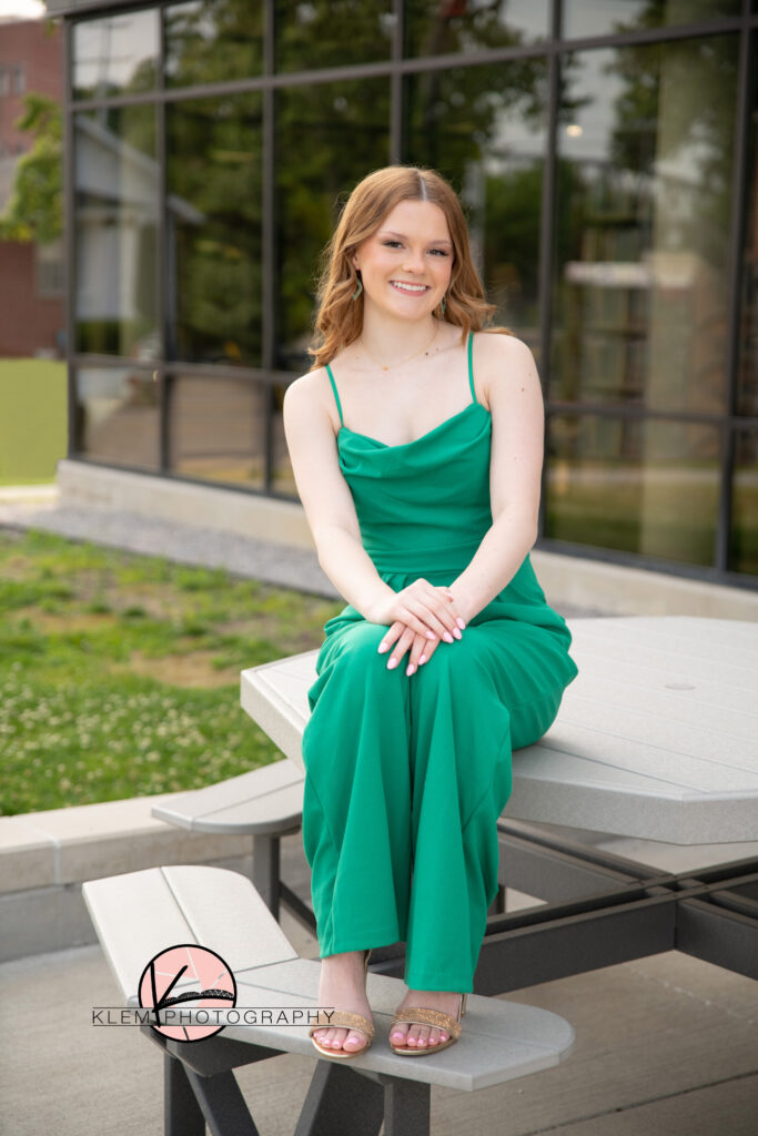 senior pictures in henderson ky by senior photographer klem photography as a senior girl sits on a table outside of the library wearing a green spaghetti strap jumpsuite with gold heels. 