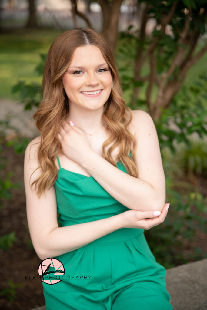 senior pictures in henderson ky by senior photographer klem photography as a senior girl sits at central park in henderson ky  wearing a green spaghetti strap jumpsuit her arms are crossed touching her neck as she smiles