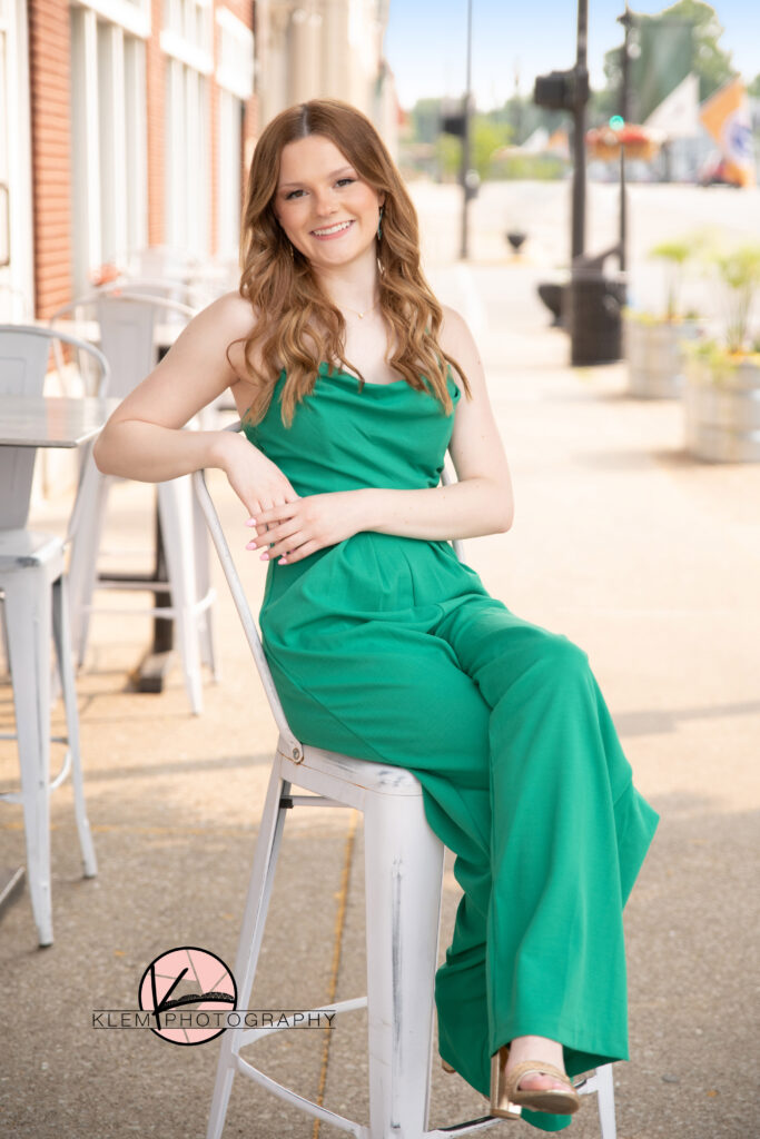 senior pictures in henderson ky by senior photographer klem photography as a senior girl sits at a bistro seat in downtown henderson at hometown roots wearing a green spaghetti strap jumpsuit with gold heels. 