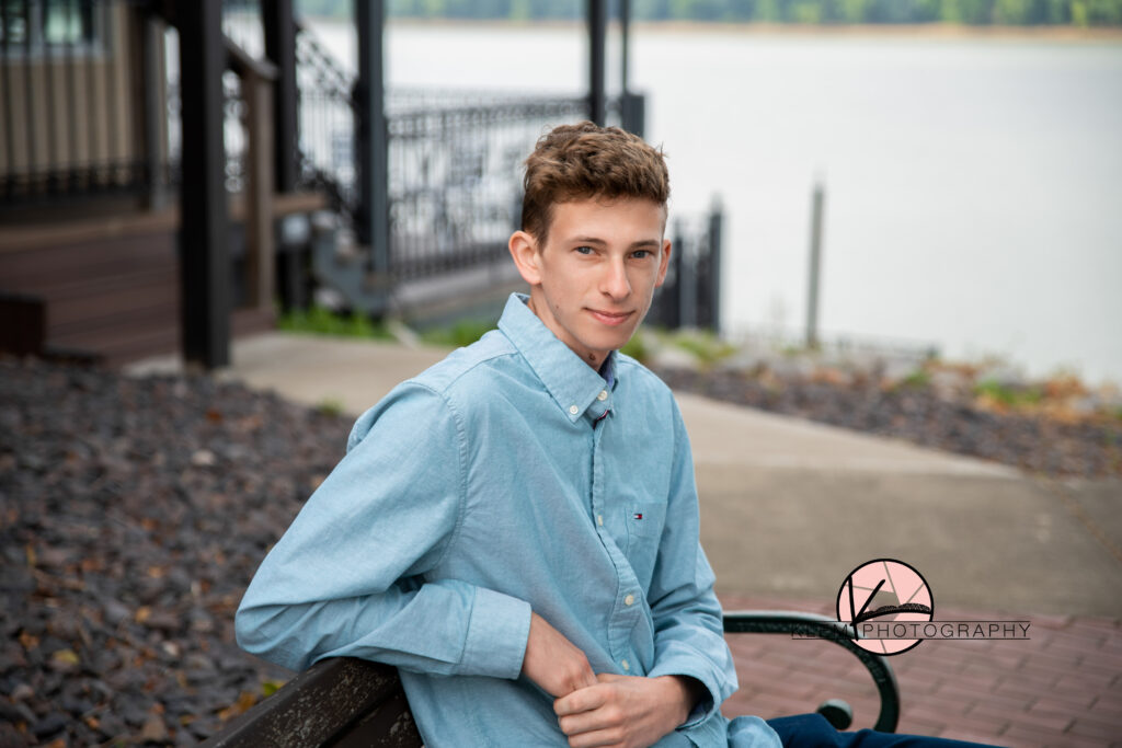 senior pictures in downtown newburgh indiana with a senior boy wearing a blue button up shirt sitting on a bench at the landing in front of the river