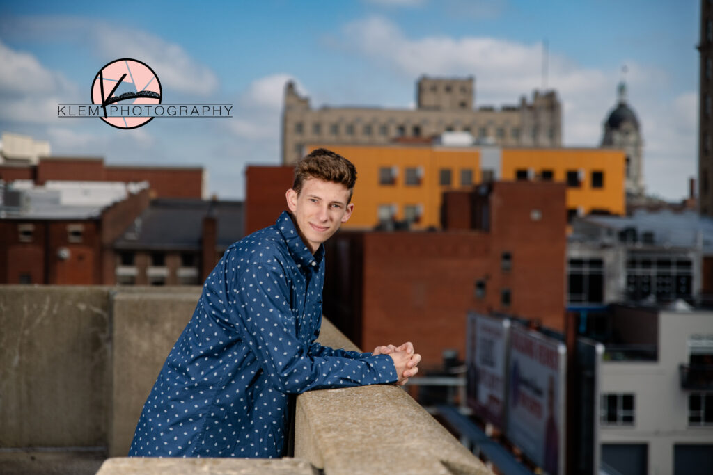 senior pictures on a rooftop in evansville indiana with the city skyline in the background. The senior boy is wearing a navy blue button up shirt with small white footballs