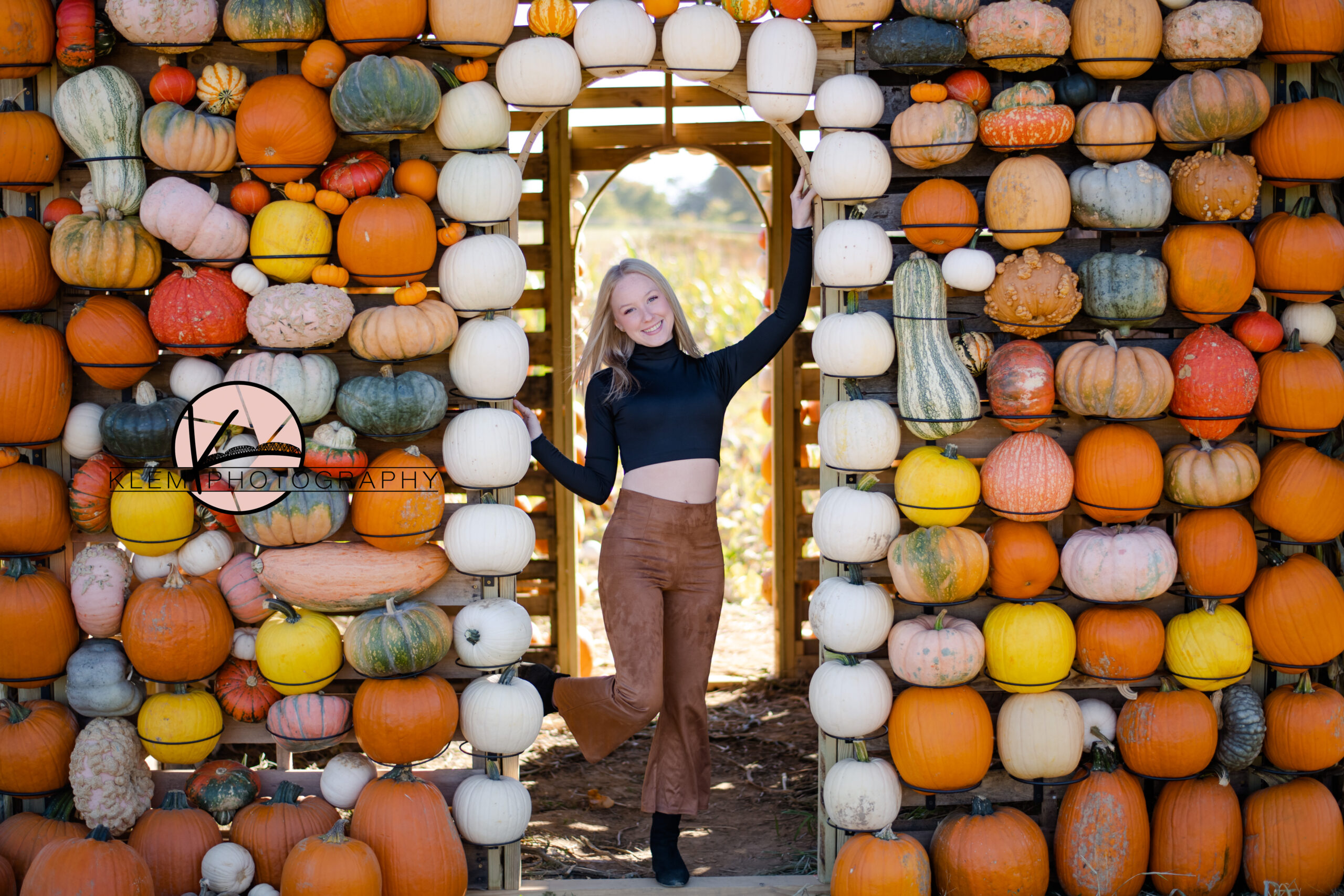 fall senior session in owensboro kentucky by kentucky senior photographer klem photography. senior girl stands in front of pumpkin wall at trunnells farm market in utica kentucky. she is wear a long sleeve black crop and brown flare pants with black boots.