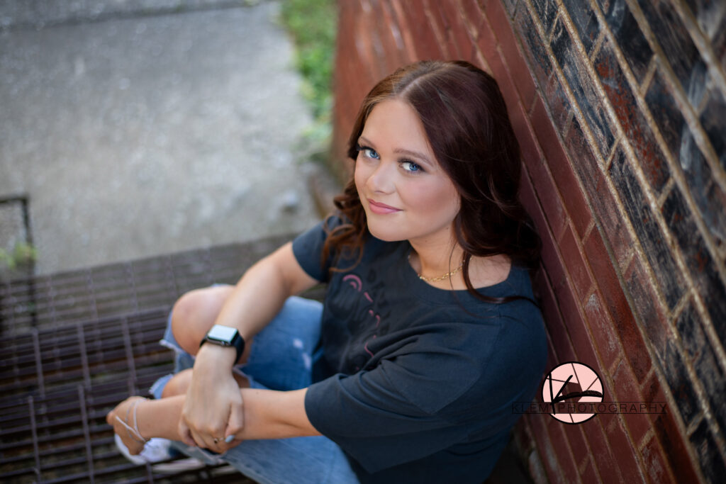henderson ky high school senior with bold blue eyes looks at the camera with a sly smile as she sits next to a rustic brick wall. 