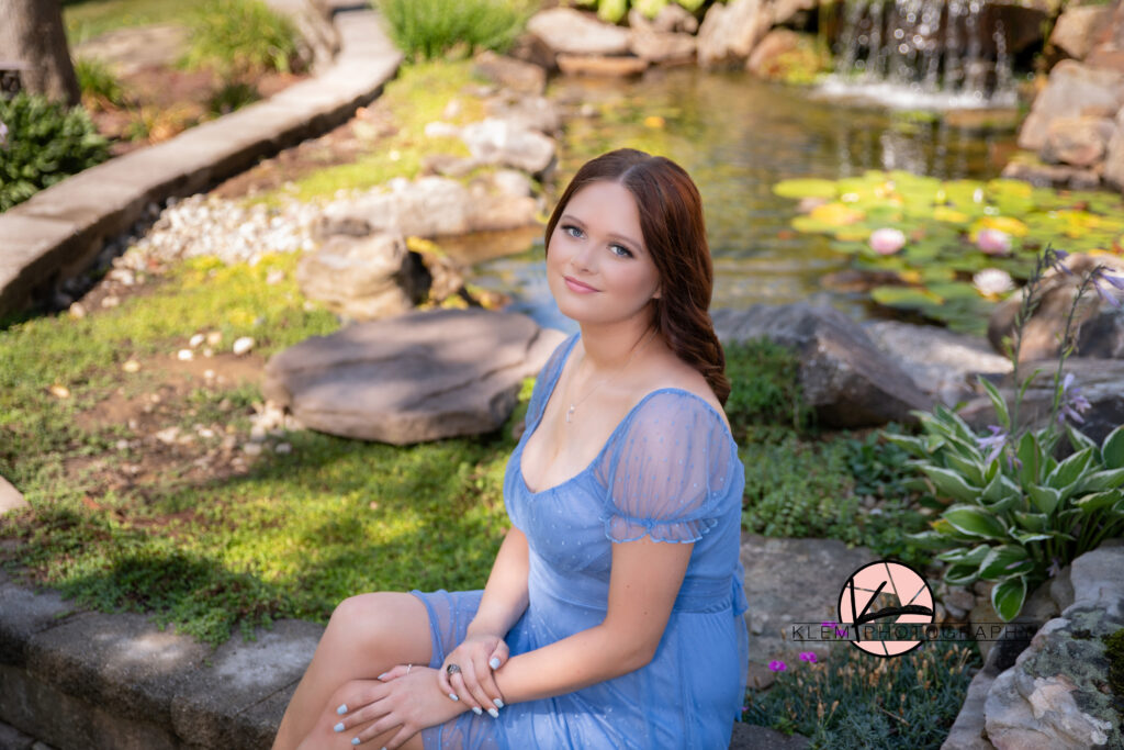 henderson ky high school senior in purple dress sits on edge of landscape water feature as her bold blue eyes and soft smile are looking at the camera