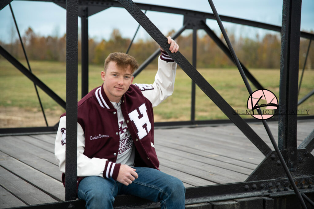Henderson County High School fall senior pictures, klem photography. senior boy wearing maroon and white letterman jacket with one hand above his head grasping the bridge railing. 