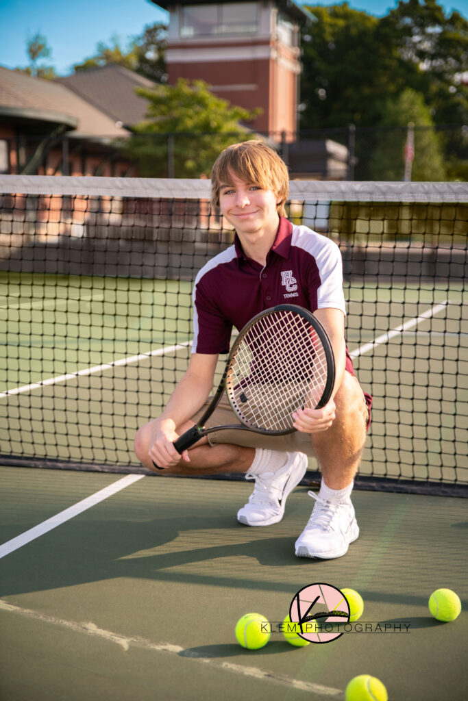 senior photos henderson ky senior boy at tennis courts in downtown henderson kentucky. he is wearing his tennis shirt from henderson county high school and its maroon and white along with khaki shorts. he is squatting and holding his tennis racket as he smiles at the camera. 