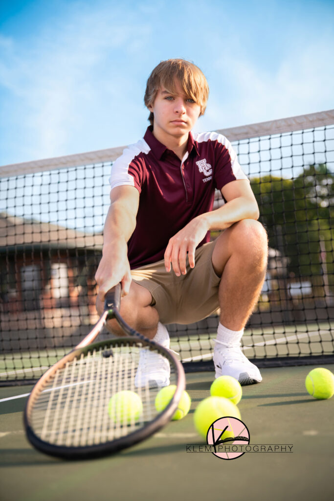 senior photos henderson ky senior boy at tennis courts in downtown henderson kentucky. he is wearing his tennis shirt from henderson county high school and its maroon and white along with khaki shorts. he is squatting and holding his tennis racket down towards the camera as he holds a straight face. 
