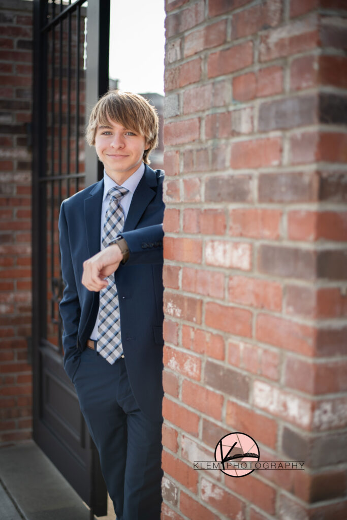 senior photos henderson ky senior boy leans with one arm against a brick wall. he is wearing a navy suit. the jacket is unbuttoned and his left hand is in his pocket. 