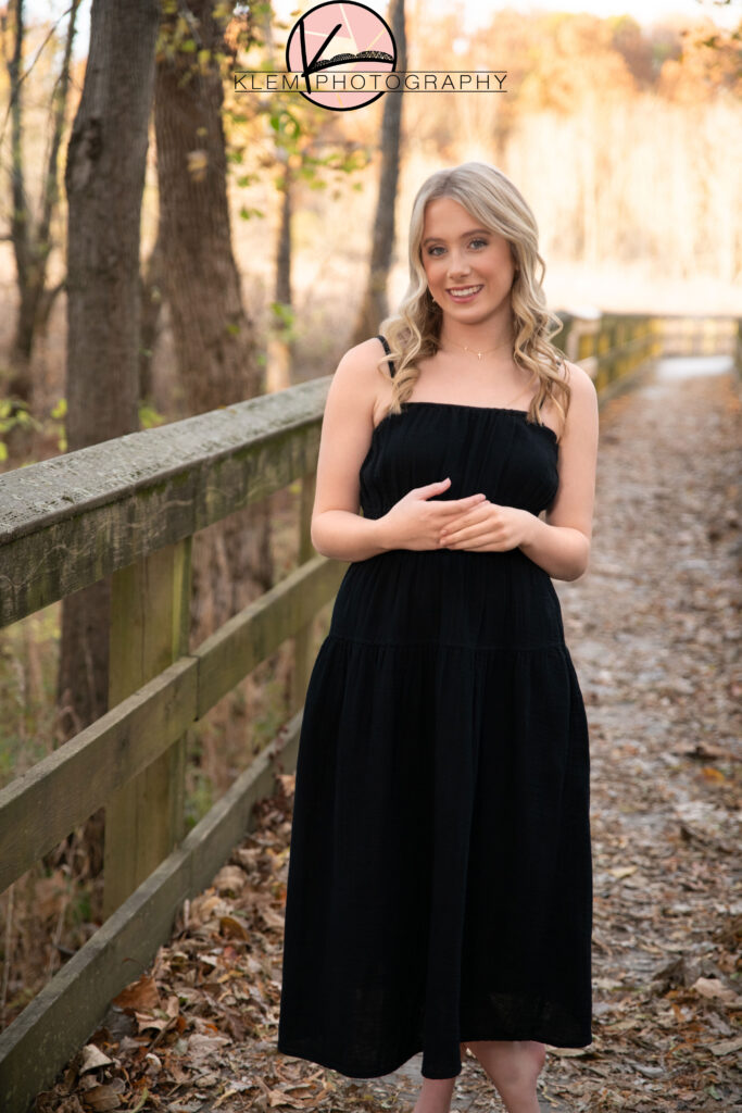 taylor swift folklore fall senior pictures by klem photography with girl in black dress standing in woods smiling at the camera by klem photography henderson ky senior photographer