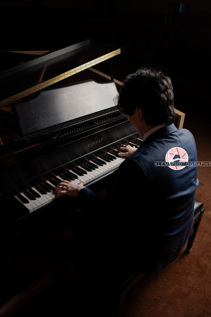Senior pictures in Henderson KY with Klem Photography at Audubon Park. Over the shoulder view of senior boy with short black hair wearing a navy blue tuxedo. He is sitting at a piano and has his hands resting on the keys.