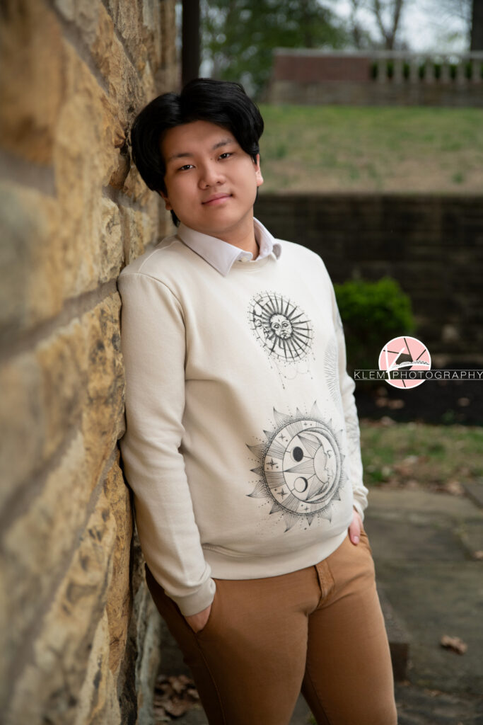 Senior pictures in Henderson KY with Klem Photography at Audubon Park. 3/4 body image of senior boy with short black hair wearing cream sweatshirt with a sun and moon on it and tan pants. He leans on a stone wall with grass in the background. His hands are in his pant pockets and he looks straight-faced at the camera.