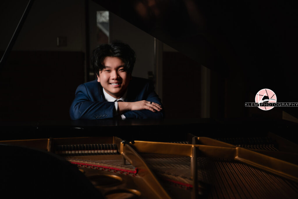 Senior pictures in Henderson KY with Klem Photography at Audubon Park. Bust view of senior boy with short black hair wearing navy blue tuxedo over a white button up. He is leaning against a piano that the inside of is visible in the foreground with his arms crossed. He is smiling at the camera.