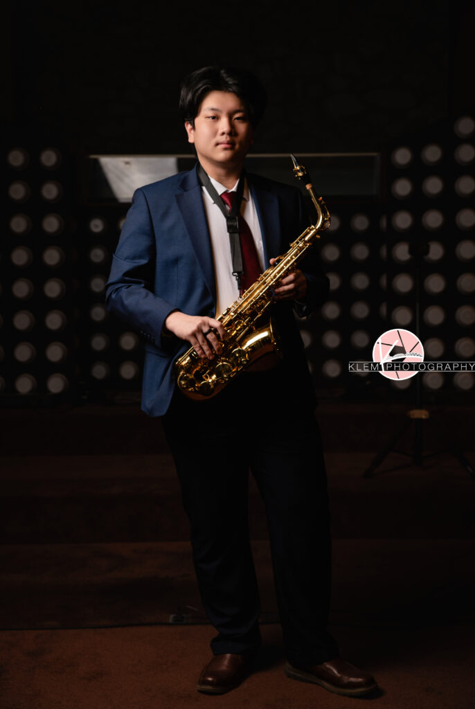 Senior pictures in Henderson KY with Klem Photography at Audubon Park. Full body view of senior boy wearing navy blue tuxedo over a white button up, red tie, and blue pants. He is holding a saxophone and looking straight-faced at the camera.