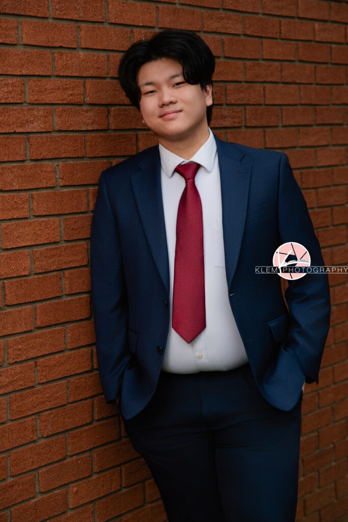 Senior pictures in Henderson KY with Klem Photography at Audubon Park. Half body view of senior boy with short black hair wearing navy blue tuxedo over a white button up and blue pants. He has his hands in his pockets and is leaning against a red brick wall as he smiles at the camera.