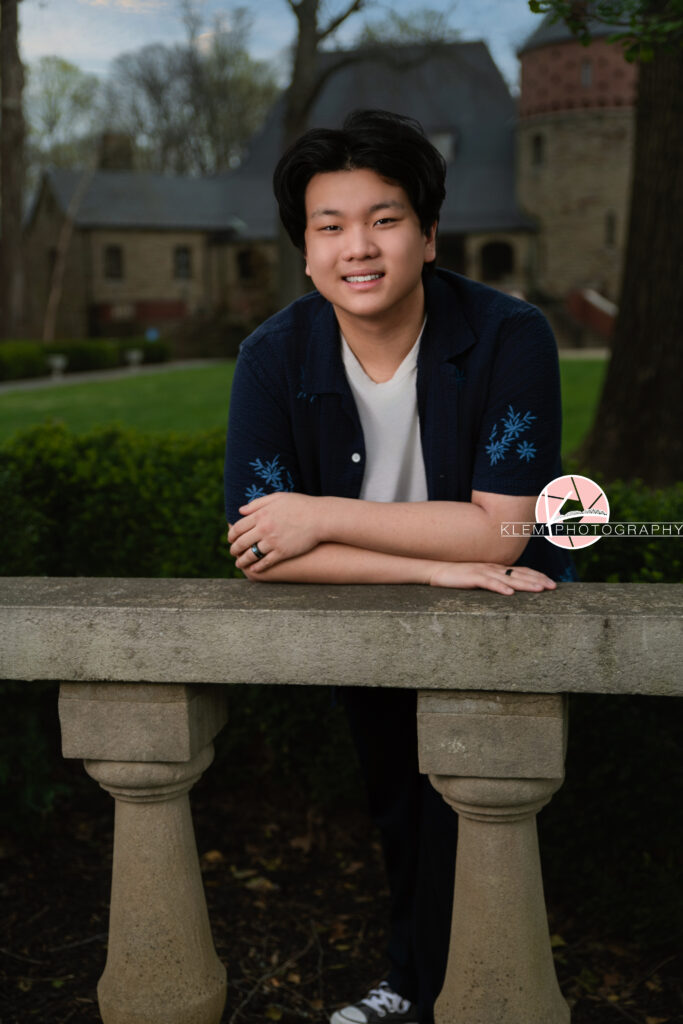 Senior pictures in Henderson KY with Klem Photography at Audubon Park. Half body image of senior boy wearing open navy button up with blue flowers over a white shirt. He is leaning up against a concrete railing, resting on his crossed arms with a castle and greenery in the background as he smiles at the camera.