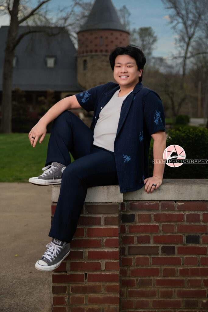 Senior pictures in Henderson KY with Klem Photography at Audubon Park. Full body image of senior boy wearing open navy button up with blue flowers over a white shirt, navy pants, and grey converse. He is sitting on a red brick wall with one leg up and an arm resting on that leg and smiling at the camera with a castle and greenery in the background.