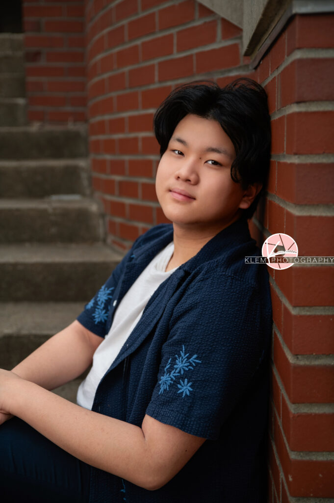 Senior pictures in Henderson KY with Klem Photography at Audubon Park. Half body image of senior boy wearing open navy button up with blue flowers over a white shirt. He is leaning up against a red brick wall and looks straight-faced at the camera with concrete stairs in the background.