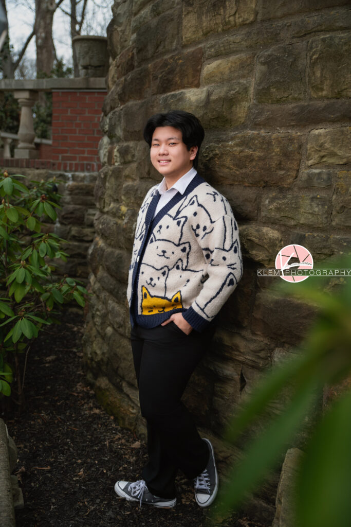 Senior pictures in Henderson KY with Klem Photography at Audubon Park. Full body image of senior boy with short black hair is wearing a cream cat cardigan and navy blue pants and grey converse. He is smiling at the camera and leaned up against a curved stone brick wall with his hands in his pant pockets.  He is standing in the dirt with bushes nearby.