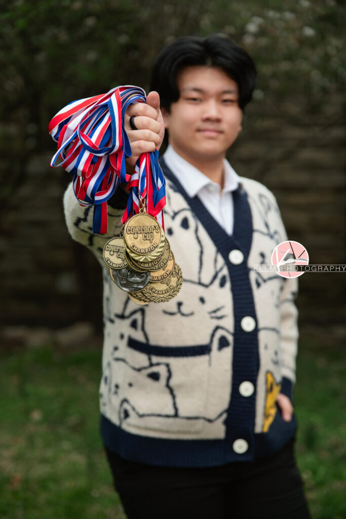 Senior pictures in Henderson KY with Klem Photography at Audubon Park. Half body image of senior boy with short black hair is wearing a cream cat cardigan and navy pants. One hand is in his cardigan pocket and the other one is holding a handful of multiple red, blue, and white medal ribbons with golden medal parts that say "governors cup." He is looking straight faced at the camera. Stone wall and grass in the background